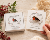 A hand holds a memorial robin brooch on a card next to its retail box on a irish made 